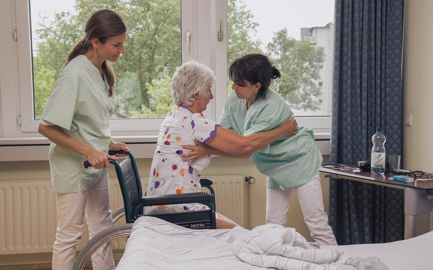 Foto: Pflegerinnen helfen älterer Frau beim Aufstehen aus dem Rollstuhl
