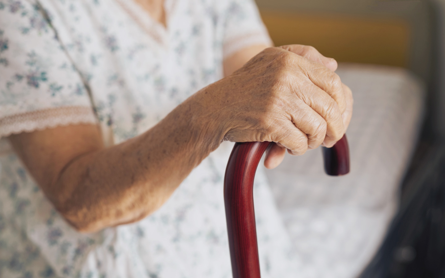Foto: Nahaufnahme der Hand einer ältere Person, sie sitzt auf einer Bettkante und hält einen Gehstock