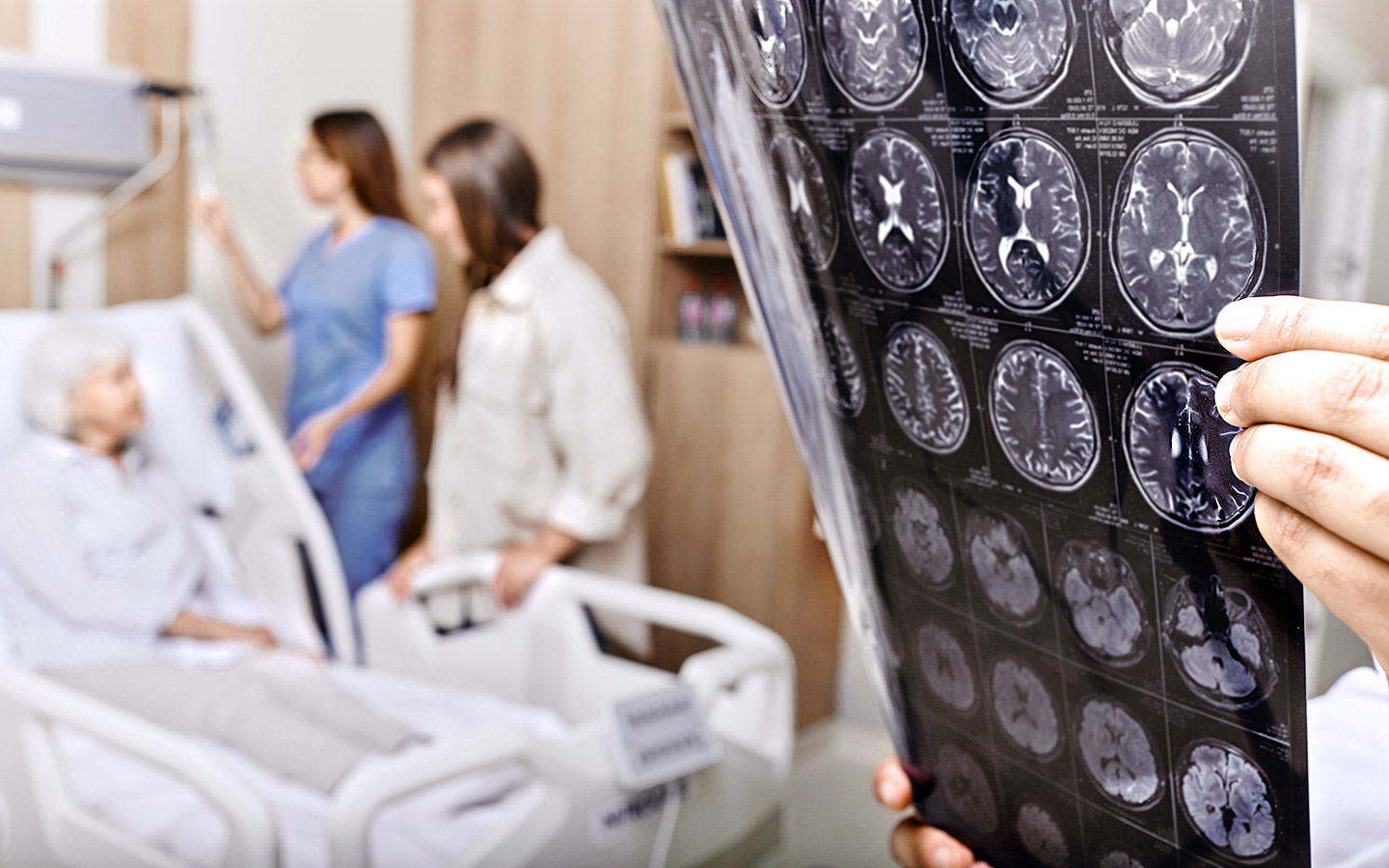 Foto: Ein Arzt hält CT-Aufnahmen von Gehirn hoch, im Hintergrund liegt eine Patientin im Krankenhausbett.